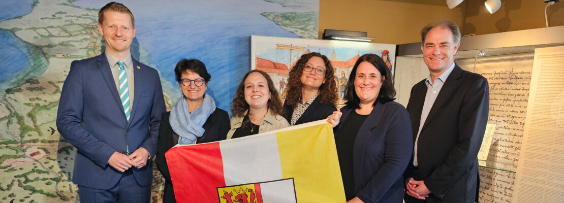 Auf dem Foto sind der Radolfzeller Oberbürgermeister, die erste Bürgermeisterin sowie das restliche Team des Stadtjubiläums Radolfzell zu sehen,, die die Flagge der Stadt Radolfzell vor sich halten.